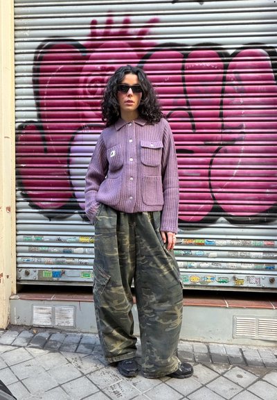 Mujer con cabello rizado oscuro que lleva gafas de sol negras, una chaqueta morada abotonada y pantalones sueltos de camuflaje, de pie contra una persiana gris con grafiti rosa.