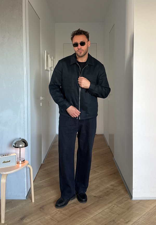 Hombre con chaqueta negra, pantalones y gafas de sol, de pie en un pasillo estrecho con suelo de madera y decoración minimalista.
