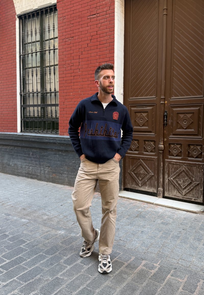 Mand iført navyblå sweater og beige bukser går på brostensbelagt gade ved siden af en rød murstensvæg og en stor trædør.