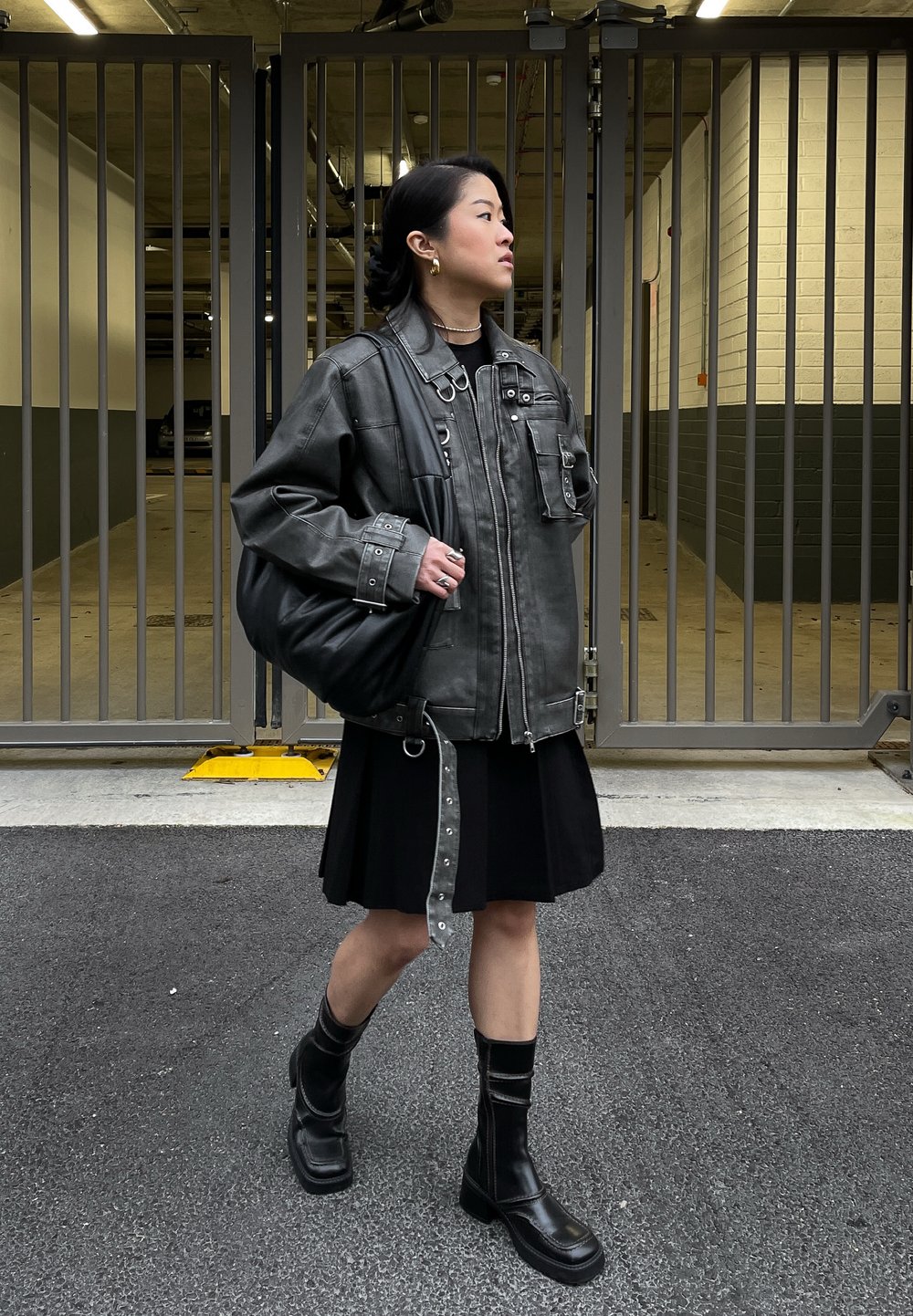 Giacca in pelle grigia oversize con molte tasche e bottoni a pressione, abbinata a un vestito nero, stivali militari neri e una borsa nera morbida.
