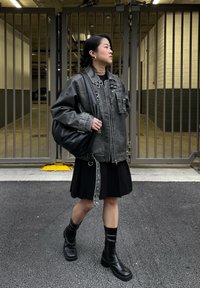 Giacca in pelle grigia oversize con molte tasche e bottoni a pressione, abbinata a un vestito nero, stivali militari neri e una borsa nera morbida.