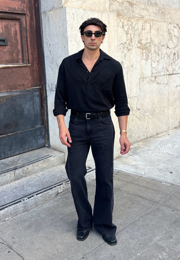 Hombre con gafas de sol negras, camisa de manga larga negra, cinturón negro, pantalones negros y zapatos negros, de pie en una acera de la ciudad cerca de un muro de piedra.