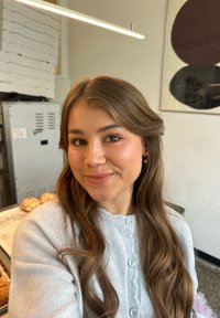 Una giovane donna con lunghi capelli marroni ondulati indossa un cardigan grigio chiaro con bottoni, sorridendo in un ambiente da caffetteria con illuminazione soffusa.