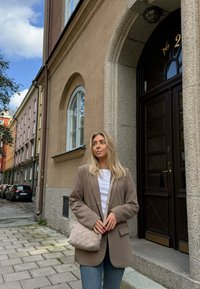 Bež blazer preko bele srajce, kombinovan s svetloplavim farmerkama. Drži tkanu svetlo bež torbu. Pozadina prikazuje smešnu kamenastu zgradu u smešnoj boji i plavo nebo.