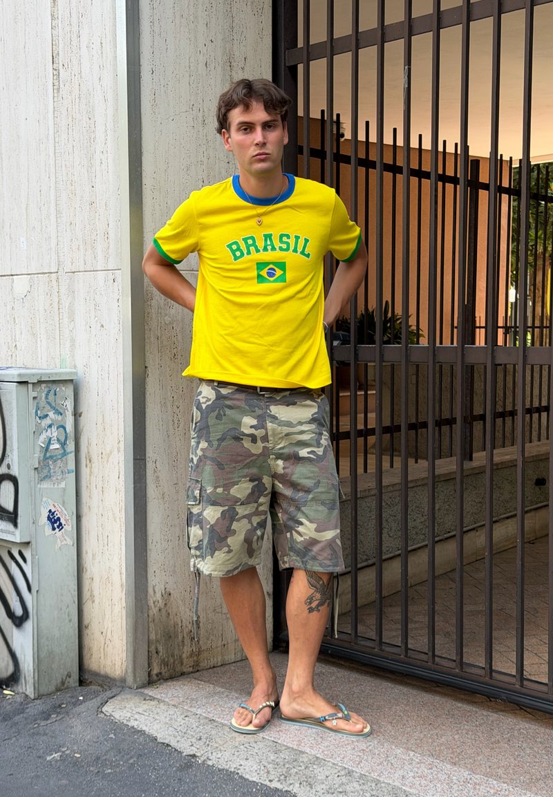 Ung mand iført en gul Brasilien t-shirt, camouflage shorts og flip-flops står med hænderne bag ryggen nær en metalport og betonvæg.