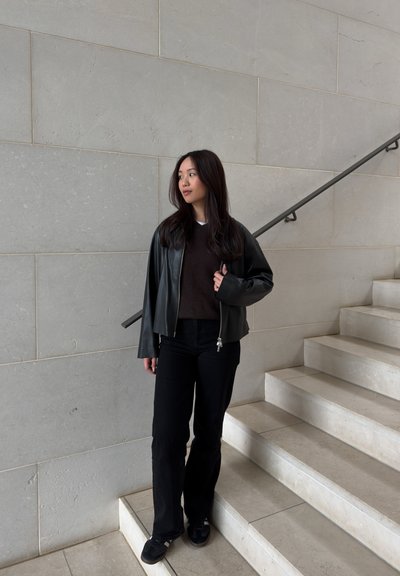 Joven mujer con cabello largo y oscuro que lleva una chaqueta de cuero negra, suéter oscuro, pantalones negros y zapatillas, parada en unas escaleras de mármol.