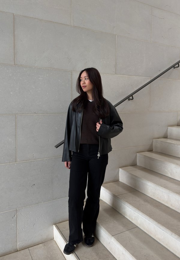 Noor naine pikkade tume juustega, kes kannab musta nahktagi, tumedat kampsunit, musti pükse ja vabaajajalatsid, seisab marmorredelitel.