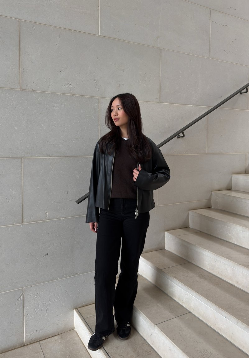 Jeune femme aux longs cheveux foncés portant une veste en cuir noire, un pull sombre, un pantalon noir et des baskets, debout sur un escalier en marbre.