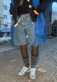 Personne portant un short en jean, des bas noirs à motifs, des baskets argentées avec "90" sur le côté, une veste noire et une écharpe bleue à franges, debout sur un trottoir urbain.