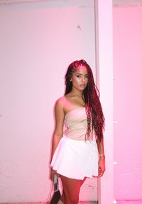 Helle beige trägerlose Bluse und weißer Minirock. Lange schwarze geflochtene Haare. Minimale Accessoires, stehend vor einem pink beleuchteten Hintergrund.