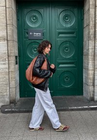 Mujer con cabello rizado que lleva una chaqueta negra, pantalones blancos y zapatillas, caminando frente a unas puertas dobles verdes con un cartel de entrada en alemán e inglés.