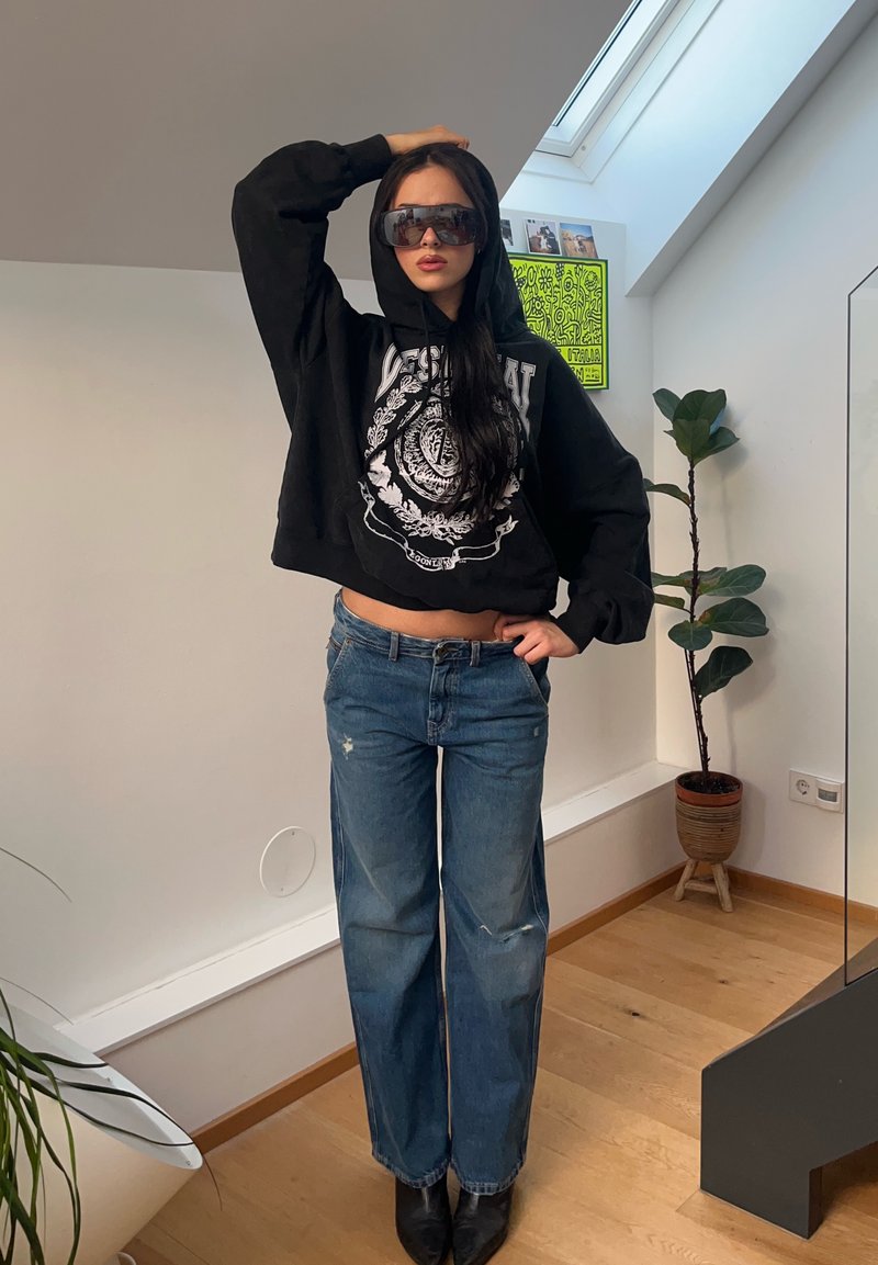 Junge Frau in übergroßem schwarzem Hoodie und Sonnenbrille posiert drinnen neben einer Pflanze, trägt lockere blaue Jeans und schwarze Stiefel.