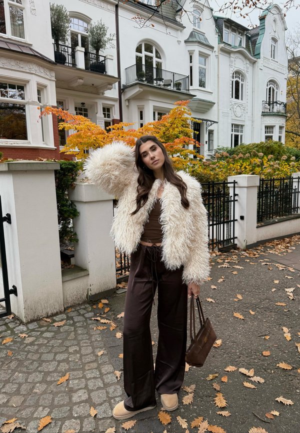 White shaggy faux fur jacket, brown top, and dark brown wide-leg pants. Beige slip-on shoes. Brown handbag. Autumn leaves in background.