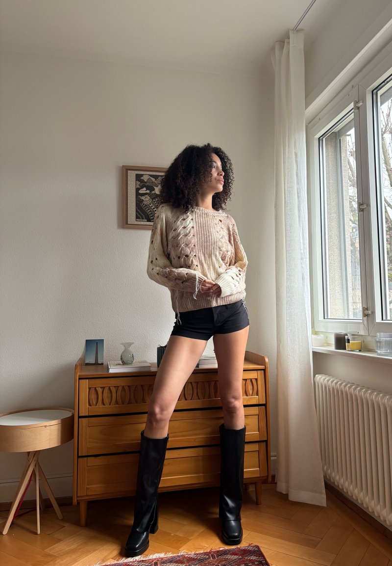Femme portant des bottes noires montantes jusqu'au genou, un short noir et un pull beige en maille torsadée, se tenant à l'intérieur près d'une fenêtre et d'une commode en bois.