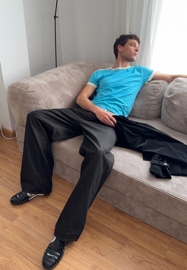 Jeune homme en t-shirt bleu et pantalon large noir se détendant sur un canapé beige en velours côtelé, regardant par une fenêtre lumineuse à droite.