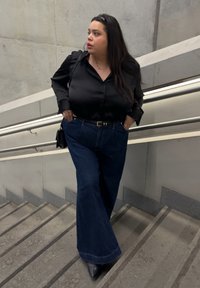 Mujer con cabello largo y oscuro, vestida con blusa negra y jeans de pierna ancha, de pie en una escalera de concreto con pasamanos de metal, mirando hacia un lado.