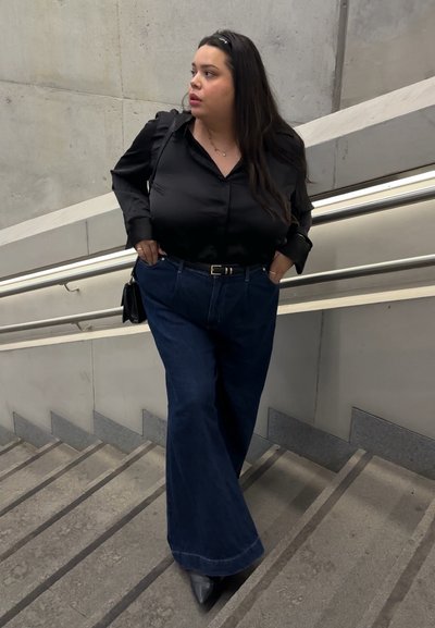 Mujer con cabello largo y oscuro, vestida con blusa negra y jeans de pierna ancha, de pie en una escalera de concreto con pasamanos de metal, mirando hacia un lado.