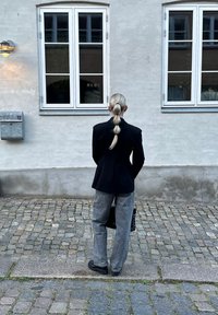 Μαύρο σακάκι κομμένο με ραφές, γκρίζο φαρδύ παντελόνι, χα hairstyle με αλογοουρά, κρατώντας μια υφή μαύρη τσάντα, στέκεται σε έναν λιθόστρωτο δρόμο δίπλα σε έναν τοίχο.