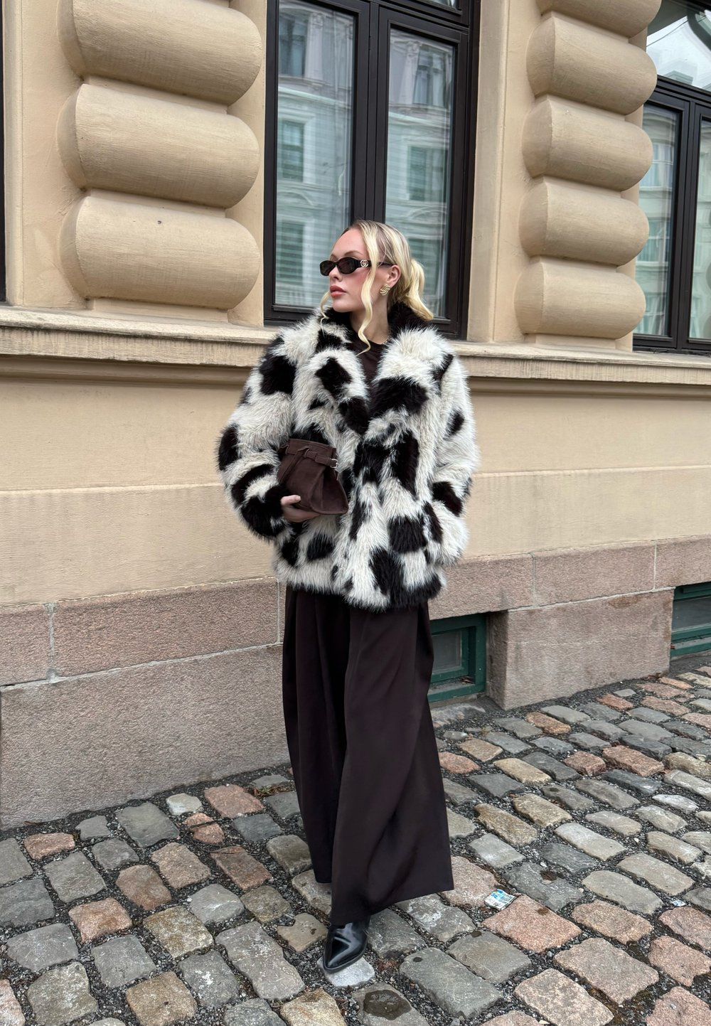 Femme debout sur une rue pavée, portant un manteau en fausse fourrure à pois noir et blanc, un pantalon large noir, des chaussures noires et des lunettes de soleil.