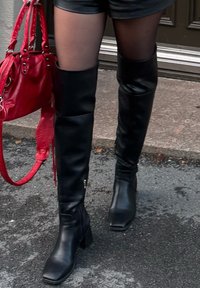 Black leather over-the-knee boots with a square toe and block heel; featuring a zip and smooth texture. Paired with black shorts and a red bag.