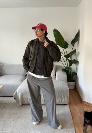 Hombre con gorra roja, chaqueta negra, suéter gris, pantalones de rayas y gafas de sol, de pie en una sala de estar con sofá, planta y un perro pequeño cerca.