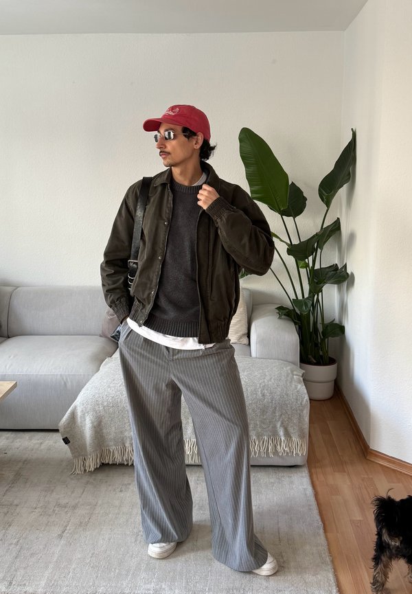 Uomo con cappellino rosso, giacca nera, maglione grigio, pantaloni a righe sottili e occhiali da sole, in piedi in un soggiorno con divano, pianta e un piccolo cane vicino.