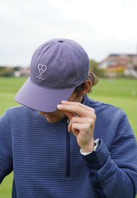 Lilla pesapalli müts tikitud südamega ja ristatud golfikeppidega logoga, kantud madalalt. Tekstureeritud tumesinine lukuga kampsun, randmel nähtav käekell.