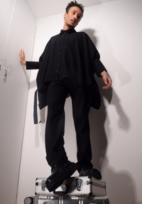 Poncho noir surdimensionné avec des boutons, associé à un pantalon noir ajusté. Le modèle se tient sur une valise métallique à la surface lisse.