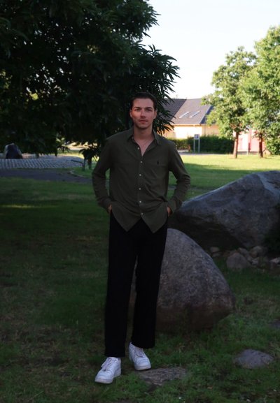 Camisa verde de manga larga, pantalones negros, zapatillas blancas. Entorno al aire libre con árboles y rocas de fondo.