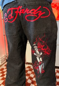 Jean noir avec broderie rouge "Ed Hardy" sur les poches arrière et un design de dague rouge et blanche avec des flammes sur la jambe droite.