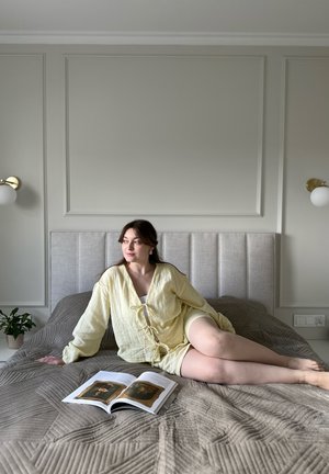 Jasnożółta, teksturowana koszula z wiązaniem z przodu, leżąca na beżowym narzucie w geometryczny wzór; na łóżku leży otwarta książka.