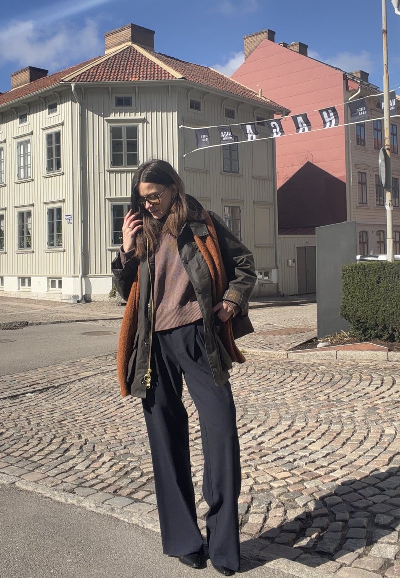 Γυναίκα με γυαλιά ηλίου, καφέ πουλόβερ, μαύρο μπουφάν και πορτοκαλί κασκόλ, που στέκεται σε μια λιθόστρωτη οδό κοντά σε ανοιχτόχρωμα κτίρια κάτω από τον ήλιο.