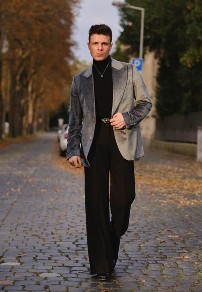Modelo vistiendo un blazer de terciopelo gris, un jersey de cuello alto negro y pantalones anchos negros, complementado con un collar de cadena y un cinturón, en un camino empedrado.