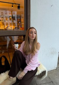 Jeune femme en pull rose et pantalon marron assise sur un banc devant un café, tenant une tasse de café à emporter et regardant sur le côté.