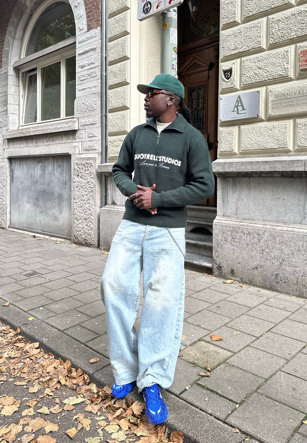 Bărbat cu șapcă verde, pulover verde închis, blugi largi bleu deschis și adidași albaștri strălucitori, stând pe un trotuar presărat cu frunze lângă o clădire de piatră.