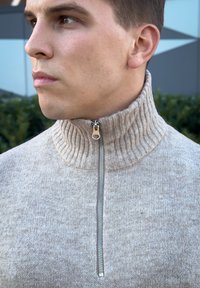 Μπεζ ζακέτα με φερμουάρ και ψηλό πλισέ γιακά, υφασμάτινο σχέδιο και ασημένια λεπτομέρεια στο φερμουάρ στο λαιμό.