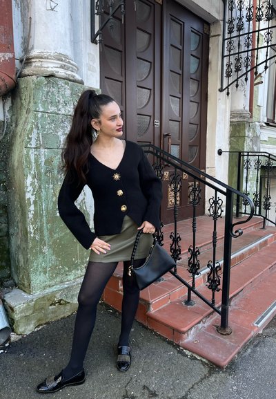 Cárdigan de punto negro con botones dorados, falda mini verde oliva, medias negras y mocasines con tachuelas. Los accesorios incluyen un bolso negro.
