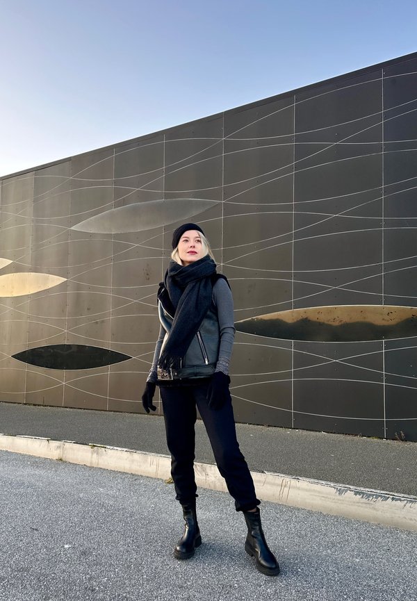 Sorte lær ankle boots, grå genser og without sleeves jakke, sort skjerf og hansker. Bakgrunnen har en moderne, teksturert svart vegg.