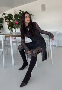 Schwarzes, durchscheinendes Langarmkleid mit Pünktchenstrumpfhosen und kniehohen, spitzen Stiefeln. Inklusive Schnürdetails und einer goldenem Kette.