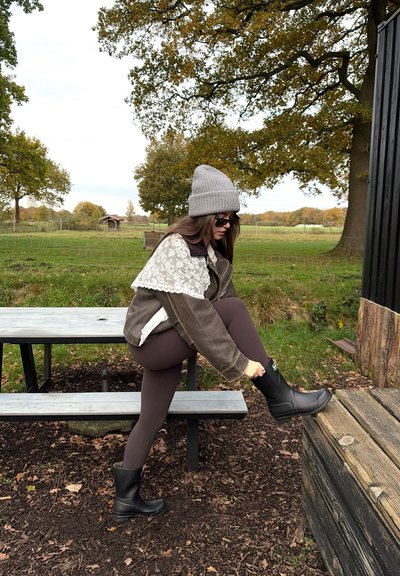 Chaqueta marrón con detalles de encaje, gorro gris, leggings negros y botas de goma negras. Ambiente exterior con césped y árboles al fondo.
