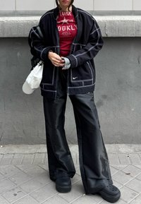 Personne debout contre un mur gris portant un pantalon noir à jambes larges, une veste noire avec des coutures blanches, un t-shirt graphique rouge, des sabots noirs, et tenant une casquette blanche.