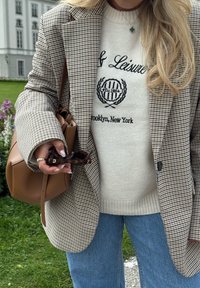 Ruskea ja beige koirankynttystakku bleiseri, valkoinen neule mustalla grafikalla, vaaleansiniset farkut, ruskea käsilaukku, pitää aurinkolaseja kädessään.