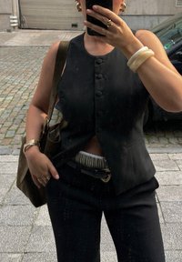 Μαύρο αμάνικο γιλέκο με κουμπιά, ψηλό κόψιμο, προσαρμοσμένος σχεδιασμός. Συνοδεύεται από μια σκούρα πράσινη τσάντα και μαύρα παντελόνια. Ορατά υλικά με υφή.