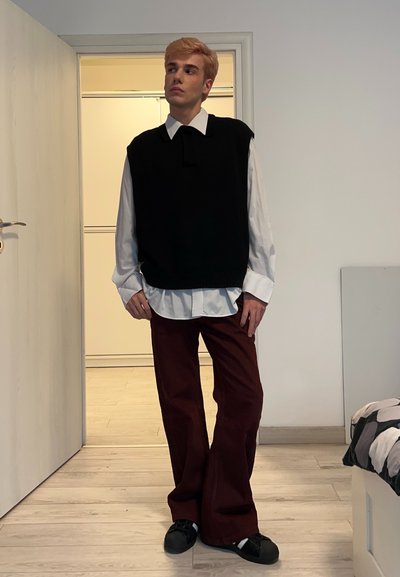 Jersey negro sin mangas sobre una camisa blanca con cuello, pantalones anchos color granate y zapatos negros. De pie en el interior con un fondo liso.