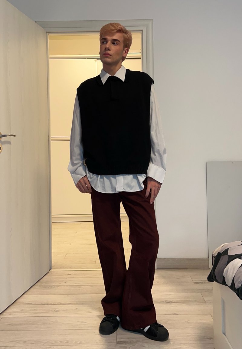 Maglione nero senza maniche sopra una camicia bianca con colletto, pantaloni ampi color marrone scuro e scarpe nere. In piedi all'interno con uno sfondo semplice.