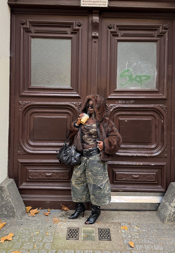 Woman wearing a camouflage top and trousers, a brown fur-lined hooded jacket, and black open-toe boots, holding a drink and a black bag, standing against large brown doors.