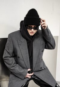 Blazer gris à double boutonnage porté sur un sweat à capuche noir, assorti à un pantalon en cuir noir brillant. Accessoires : lunettes de soleil noires et bonnet en maille.