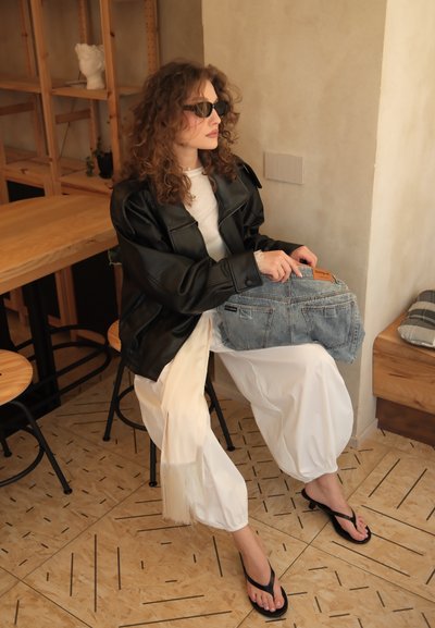 Mujer con cabello rizado que lleva gafas de sol, chaqueta de cuero negra, pantalones blancos y chanclas, sentada dentro de una habitación sosteniendo un bolso vaquero en su regazo.