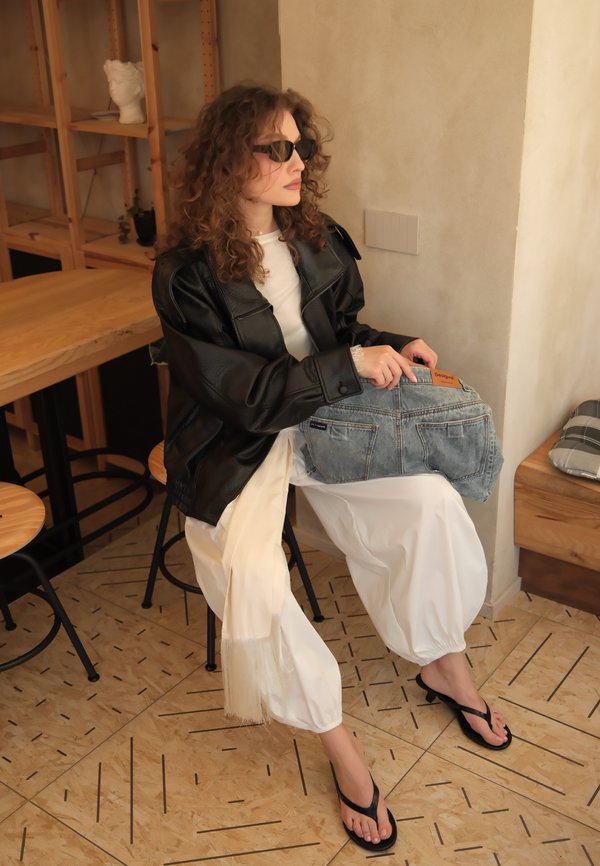 Femme aux cheveux bouclés portant des lunettes de soleil, une veste en cuir noire, un pantalon blanc et des tongs, assise à l'intérieur en tenant un sac en jean sur ses genoux.