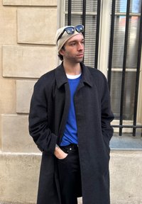 Άνδρας που φορά μαύρο πανωφόρι, μπλε πουλόβερ, μπεζ σκουφάκι με γυαλιά ηλίου στην κορυφή, στέκεται με τα χέρια στις τσέπες κοντά σε πέτρινο τοίχο και παράθυρο με κάγκελα.